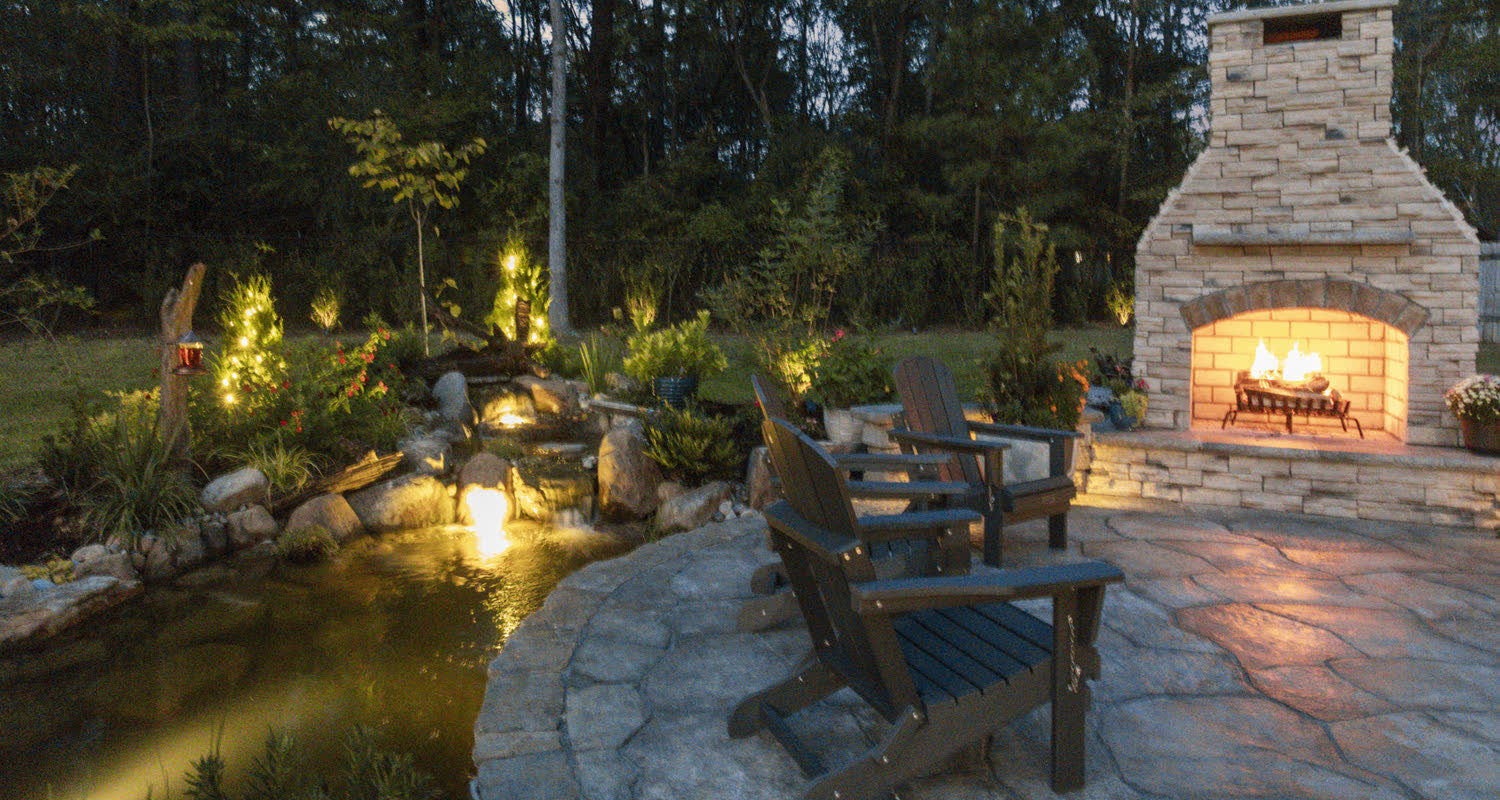 A Superscapes backyard pond and stream beside a stone fireplace and seating at dusk